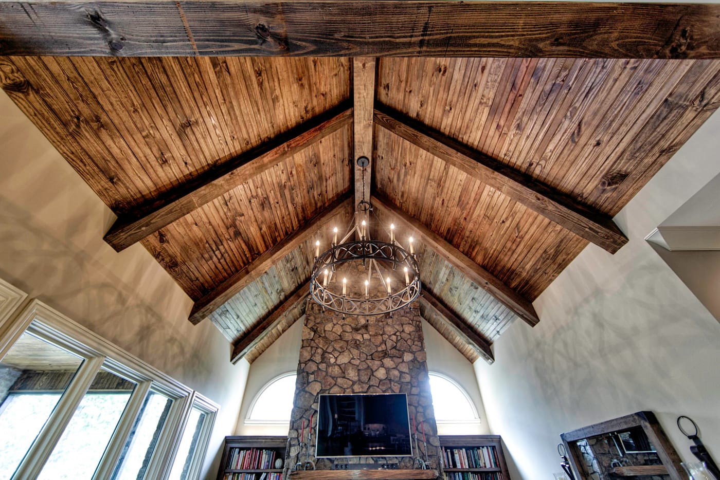 Looking up at a vaulted wood-plank ceiling with exposed beams, a round iron chandelier and a stone fireplace