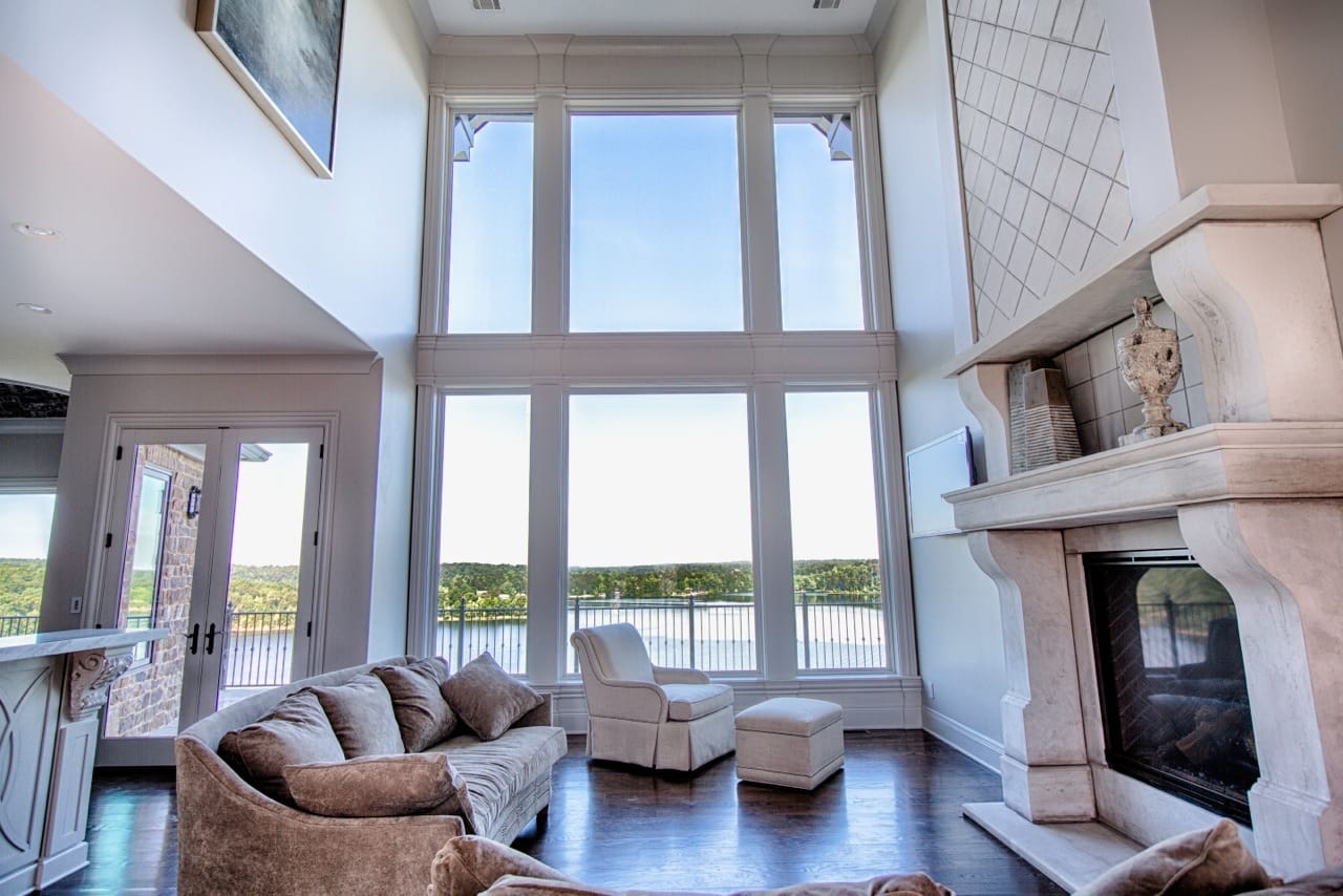 Two-story living room with a wall of tall windows overlooking the water, a carved stone fireplace and neutral seating