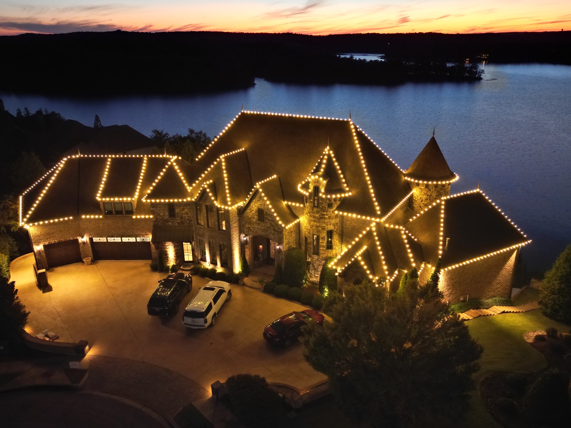 Aerial front view of a stone and brick lakeside home at sunset, with turrets, a curved driveway and lights tracing the rooflines