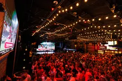 Wide view of the completed Two Dimes main room with ceiling lights, screens, raised seating and an event crowd